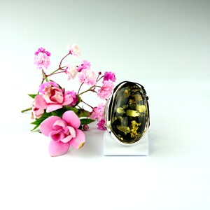 May include: A silver ring with a large, oval-shaped green amber stone. The stone has a dark green color with flecks of gold throughout. The ring is set on a white stand against a white background.
