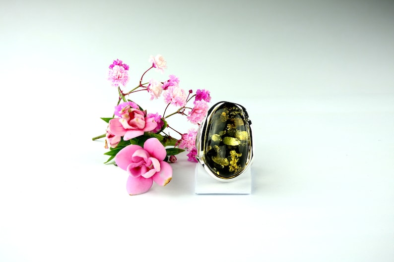 May include: A silver ring with a large, oval-shaped green amber stone. The stone has a dark green color with flecks of gold throughout. The ring is set on a white stand against a white background.