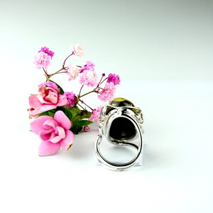 May include: A silver ring with a large, round, dark stone set in a decorative, ornate setting. The ring is displayed on a white surface with pink flowers.