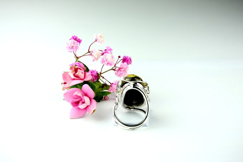 May include: A silver ring with a large, round, dark stone set in a decorative, ornate setting. The ring is displayed on a white surface with pink flowers.