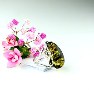 May include: A silver ring with a large oval green amber stone. The stone has a swirling pattern of gold and brown. The ring is displayed on a white surface with pink flowers.