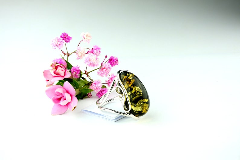 May include: A silver ring with a large oval green amber stone. The stone has a swirling pattern of gold and brown. The ring is displayed on a white surface with pink flowers.