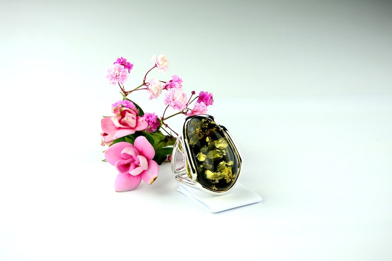 May include: A silver ring with a large, oval-shaped green amber stone. The stone has a unique, marbled pattern and is set in a simple, elegant band. The ring is surrounded by pink flowers.
