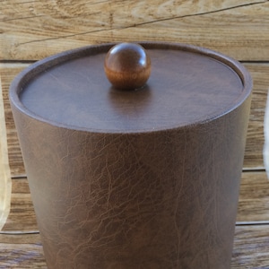 Puede incluir: Un cubo de hielo de cuero marrón con una tapa de madera y una perilla redonda de madera en la parte superior.