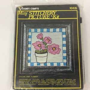 May include: A mini embroidery kit with a black frame and a white mat with a blue and white checkered border. The design is of pink flowers in a white pot. The kit includes 100% acrylic crewel yarns, an embroidery needle, and an illustrated stitch chart. The kit is 5 inches by 5 inches and fits a 4 inch by 4 inch frame.