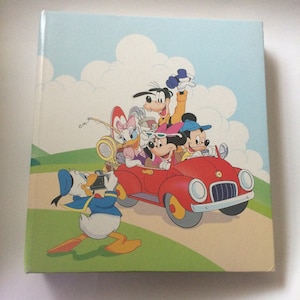 May include: A square, hardback book with a vibrant illustration of Disney characters. Mickey Mouse drives a red car with Goofy, Minnie Mouse, and Daisy Duck. Donald Duck takes a photo. The background shows a blue sky with white clouds and green hills.