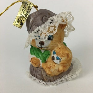 May include: A ceramic ornament of a brown bear wearing a gray hat with white lace trim, holding a smaller brown bear. The ornament has a gold tag that reads "Caring Critter Chimers TM".