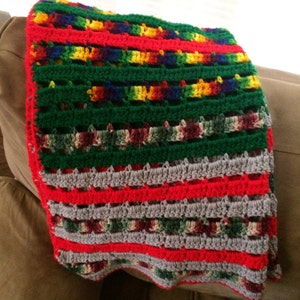 Puede incluir: Una manta de crochet colorida con rayas de hilo rojo, verde, gris y arcoíris. La manta está doblada y drapeada sobre un sofá marrón.