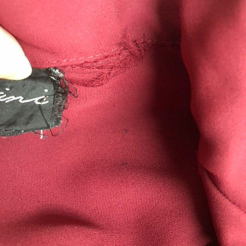 K&ouml;nnte beinhalten: Nahaufnahme eines burgunderfarbenen Stoffes mit einem schwarzen Etikett, auf dem "Giambattista Valli" steht. Der Stoff hat einen kleinen Riss.