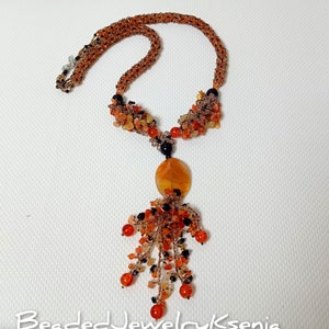 May include: An orange beaded necklace with a pendant. The necklace features a woven design with orange and black beads. The pendant has an oval orange stone with a cluster of orange and black beads hanging below. The text "BeadedJewelryKsenia" is at the bottom.