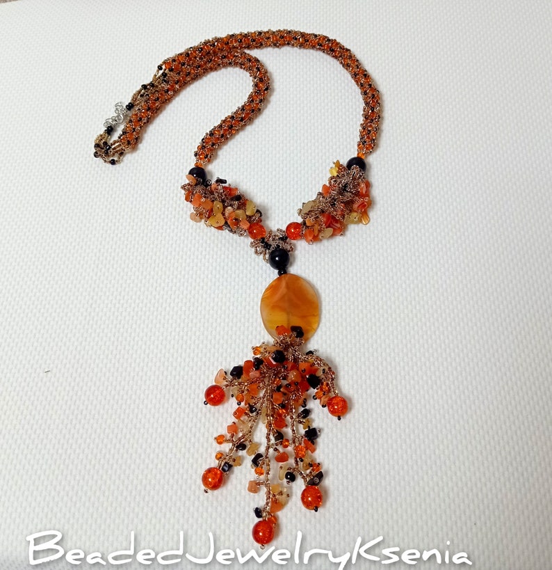 May include: An orange beaded necklace with a pendant. The necklace features a woven design with orange and black beads. The pendant has an oval orange stone with a cluster of orange and black beads hanging below. The text "BeadedJewelryKsenia" is at the bottom.