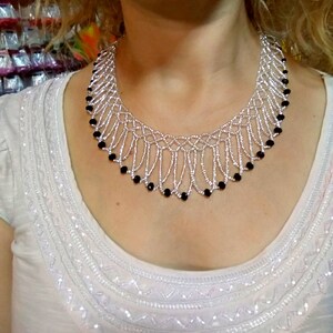 May include: A silver beaded necklace with a delicate, openwork design. The necklace features strands of silver beads and is accented with small, black beads along the bottom edge. The necklace is worn on a person wearing a white top.