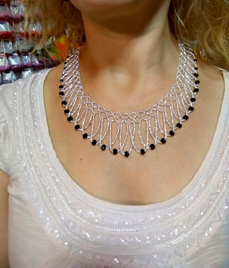 May include: A silver beaded necklace with a delicate, openwork design. The necklace features strands of silver beads and is accented with small, black beads along the bottom edge. The necklace is worn on a person wearing a white top.