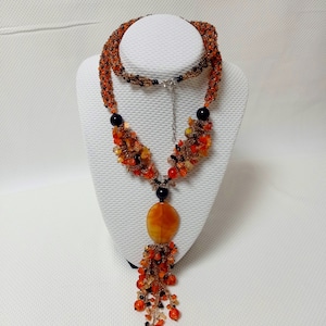 May include: A handcrafted beaded necklace with orange, black, and amber-colored beads. The necklace features a large, oval amber pendant and a cascading fringe of beads. The necklace is displayed on a white bust.