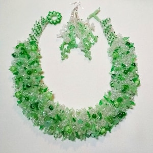May include: A beaded jewelry set featuring a necklace and earrings. The necklace and earrings are made of clear and green beads. The necklace is a collar style, and the earrings are dangling. The set is displayed on a white surface.