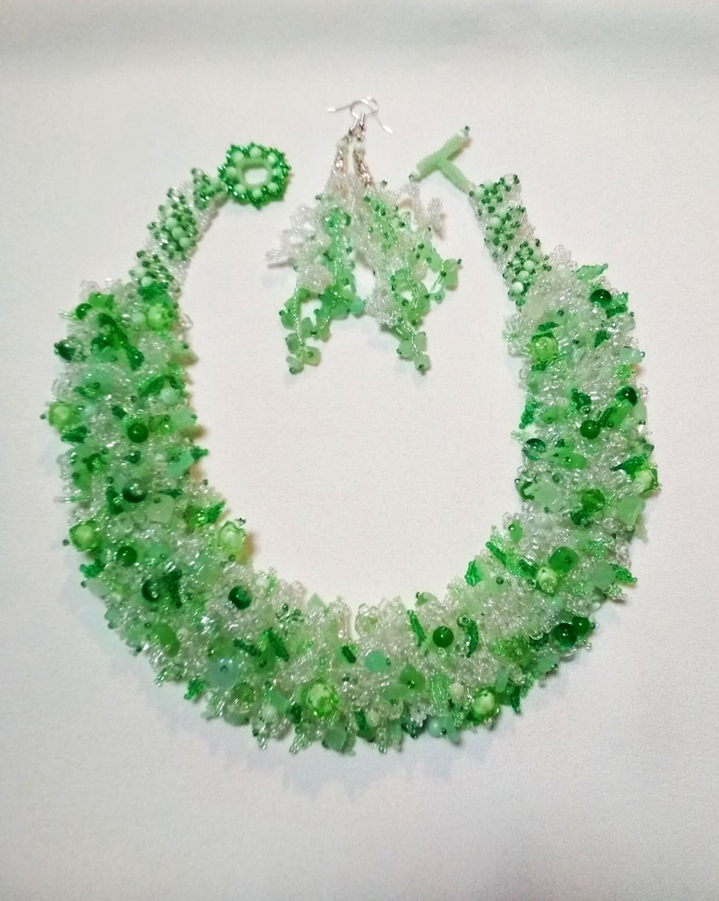 May include: A beaded jewelry set featuring a necklace and earrings. The necklace and earrings are made of clear and green beads. The necklace is a collar style, and the earrings are dangling. The set is displayed on a white surface.