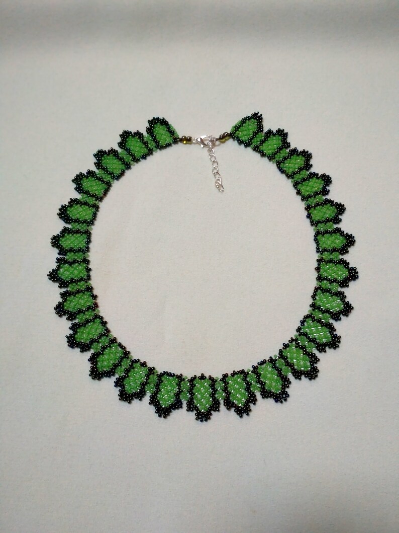 May include: A beaded necklace with a repeating leaf design. The leaves are bright green with black beaded borders. The necklace has a silver clasp and is shown against a pale background.