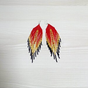May include: A pair of beaded earrings with a feather-like design. The earrings feature a gradient of colors, starting with red at the top, transitioning to gold, and ending with black at the bottom. The earrings are displayed on a white wooden surface.