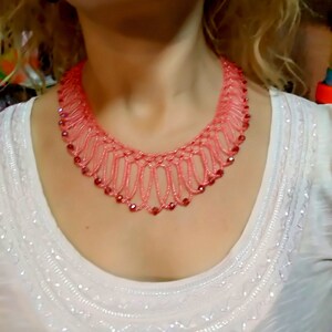 May include: A coral-colored beaded necklace with a delicate, openwork design. The necklace features small, round red beads and a scalloped edge. The necklace is worn over a white top with embroidered details.