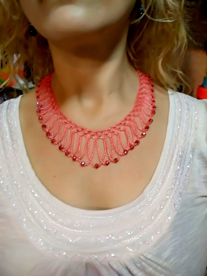 May include: A coral-colored beaded necklace with a delicate, openwork design. The necklace features small, round red beads and a scalloped edge. The necklace is worn over a white top with embroidered details.