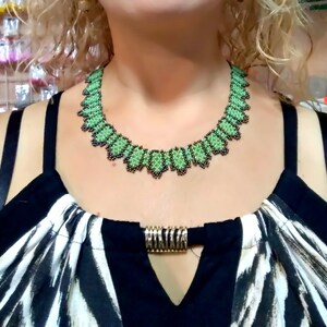 May include: A green and black beaded necklace with a scalloped edge. The necklace is worn over a black and white patterned top, with a gold-coloured clasp.