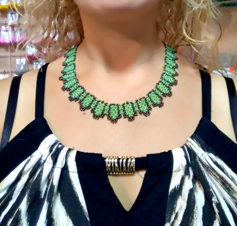 May include: A green and black beaded necklace with a scalloped edge. The necklace is worn over a black and white patterned top, with a gold-coloured clasp.