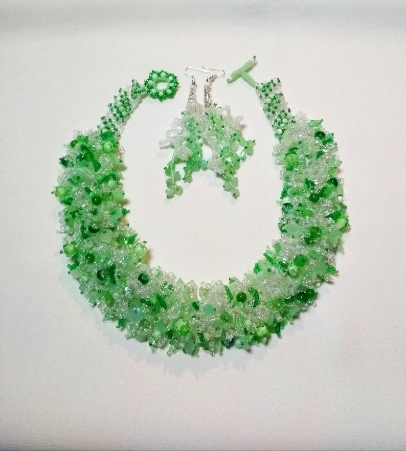May include: A beaded jewelry set featuring a necklace, earrings, and a ring. The set is composed of clear and green beads, creating a textured, organic appearance. The necklace has a toggle clasp. The earrings have silver hooks.