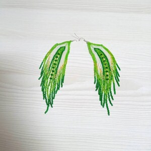 May include: A pair of handmade beaded earrings in a feather design. The earrings feature a gradient of green beads, with white and gold accents. The fringe-style earrings have silver-colored ear wires.