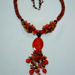 May include: A beaded necklace in shades of orange, red, and black. The necklace features a heart-shaped design with a large oval pendant and a cluster of dangling beads. The necklace is made of small beads and has a silver clasp.
