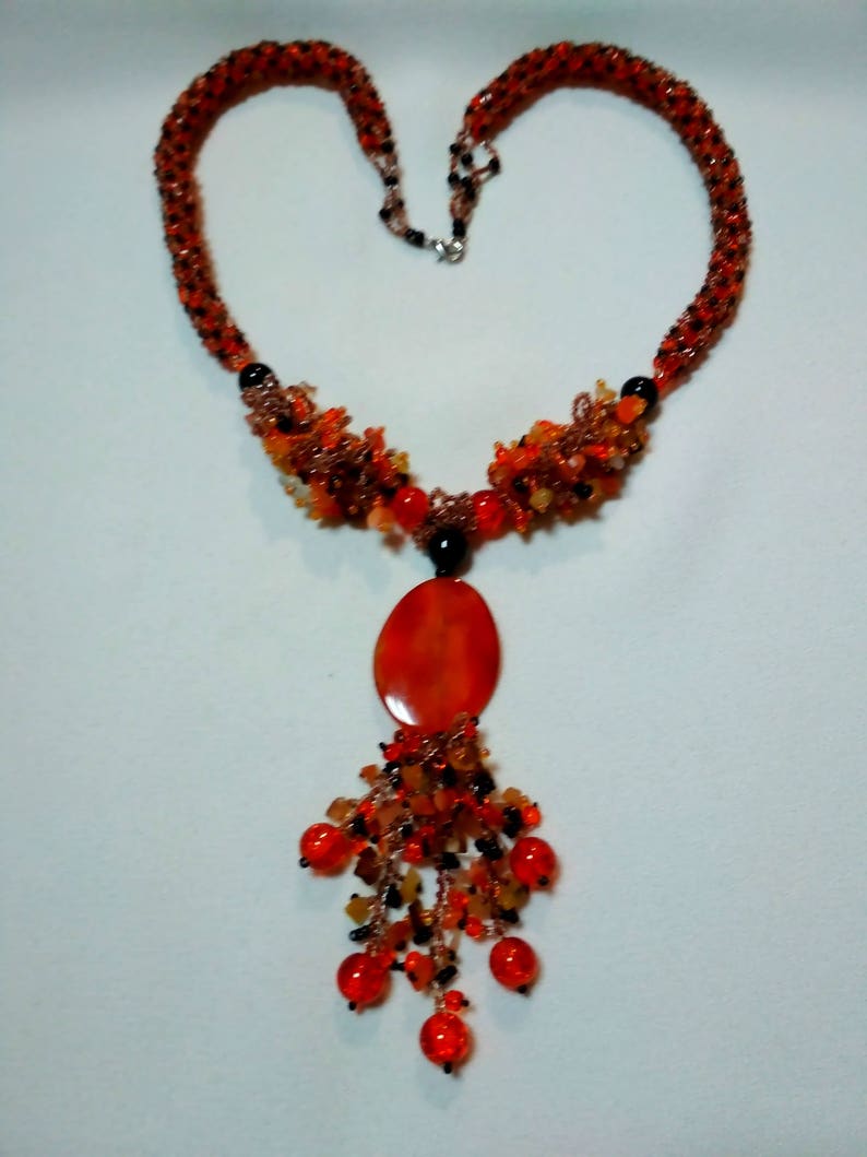 May include: A beaded necklace in shades of orange, red, and black. The necklace features a heart-shaped design with a large oval pendant and a cluster of dangling beads. The necklace is made of small beads and has a silver clasp.