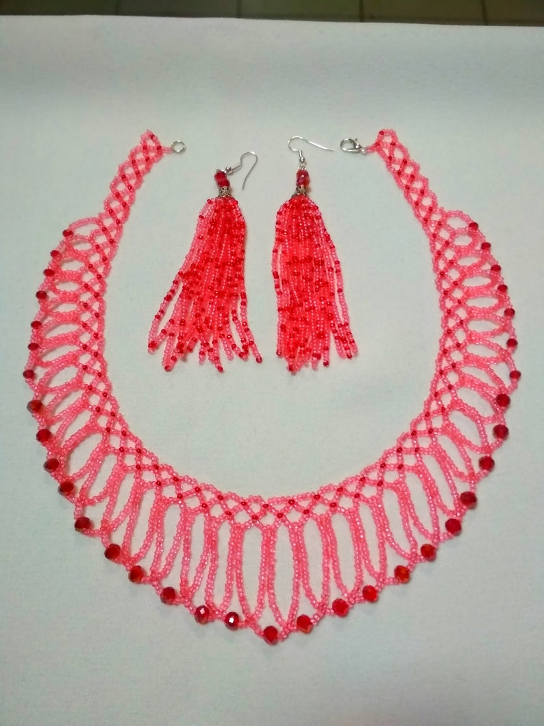 May include: A pink beaded necklace and matching earrings set. The necklace features a delicate, openwork design with red accents. The earrings have long, red beaded tassels, creating a coordinated jewelry set.
