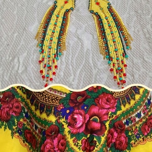 May include: A pair of handmade beaded earrings with a yellow base and cascading strands of yellow, brown, black, turquoise, and red beads. The earrings are displayed on a textured white surface. A yellow floral scarf with red roses is in the background.