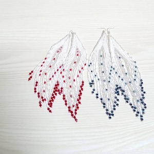 May include: Two pairs of beaded earrings. One pair features white beads with red accents, the other with white and dark blue beads. The earrings have a long, fringed design, with silver-colored metal hooks.