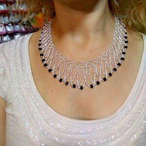 May include: A silver beaded necklace with a fringe design. The necklace features small silver beads and is accented with black beads. The necklace is worn on a white top.