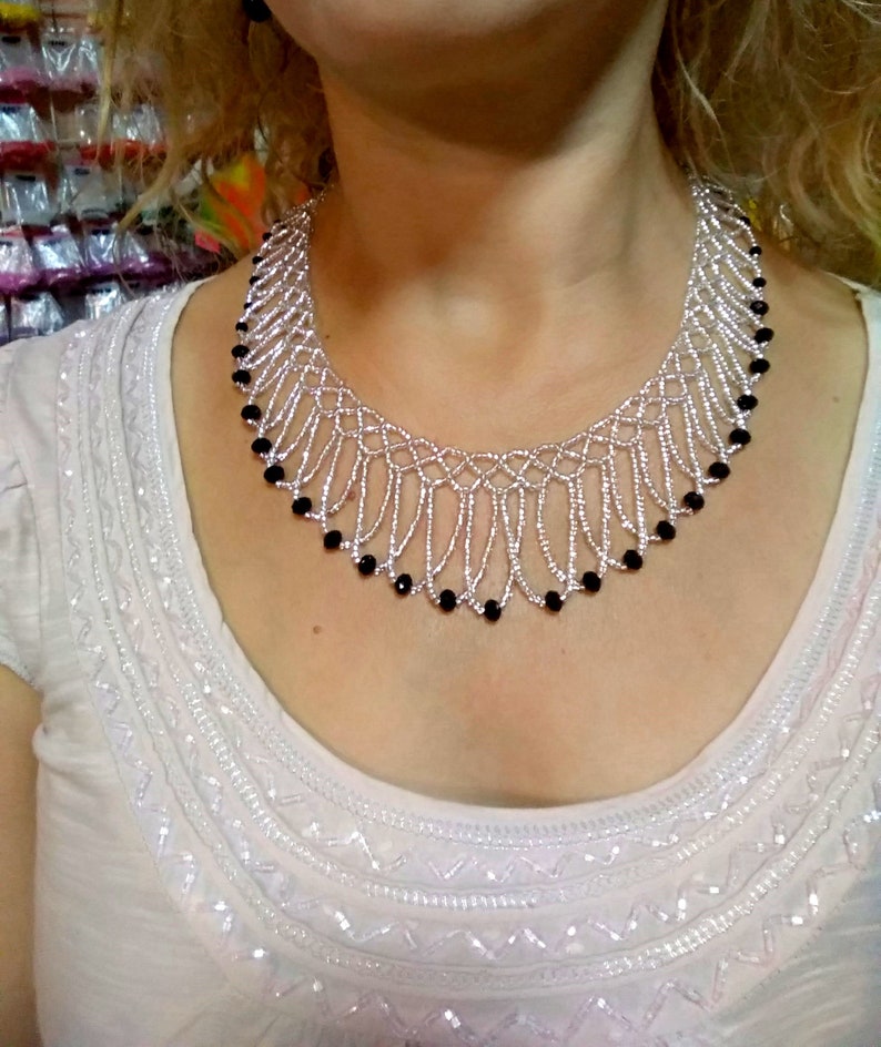 May include: A silver beaded necklace with a fringe design. The necklace features small silver beads and is accented with black beads. The necklace is worn on a white top.