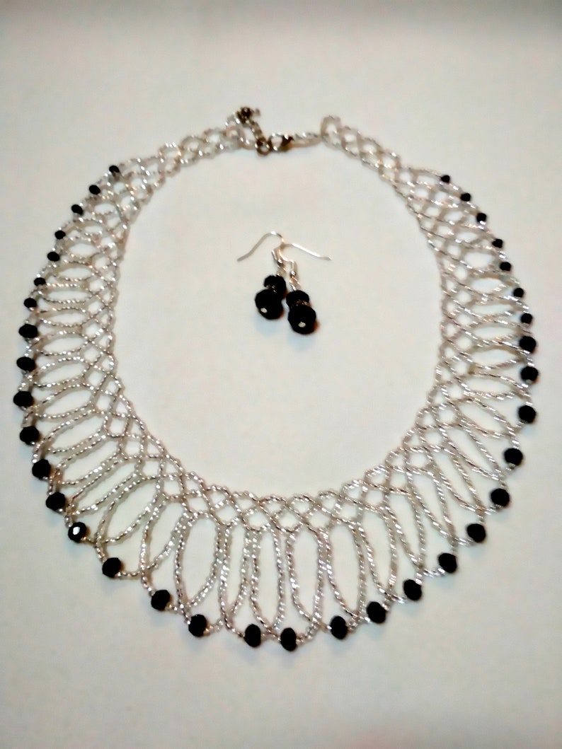 May include: A silver-toned necklace and matching earrings set. The necklace features a woven design with black beads. The earrings have black beads. The jewelry set is displayed on a white surface.