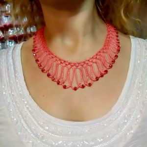 May include: A coral-colored beaded necklace with a delicate, openwork design. The necklace features small, red beads interspersed throughout the coral strands. The necklace is worn over a white, embellished top.