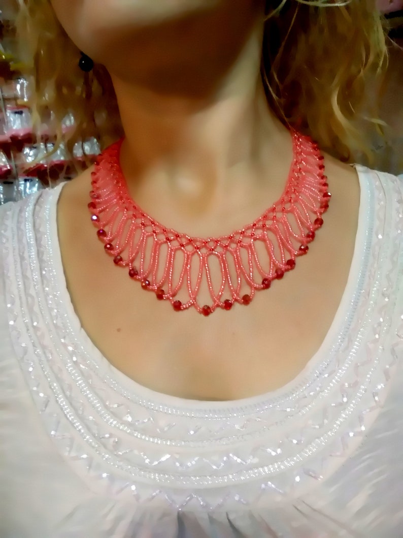 May include: A coral-colored beaded necklace with a delicate, openwork design. The necklace features small, red beads interspersed throughout the coral strands. The necklace is worn over a white, embellished top.