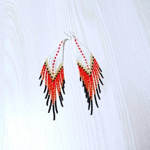 May include: A pair of beaded earrings with a white, red, black, and gold color scheme. The earrings have a long, fringed design, with a white top section, transitioning to red, and ending with black beads. The earrings are made with small beads.
