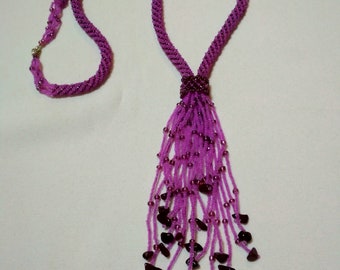 May include: A handmade purple beaded necklace with a twisted rope design. The necklace features a tassel of smaller beads and dark purple stone accents. The necklace is secured with a silver clasp. The jewelry is displayed on a white background.