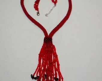 May include: A red beaded necklace with a heart-shaped design. The necklace features a woven beaded collar that splits into a beaded fringe with dark red accents. The necklace has a silver-colored clasp and adjustable chain.