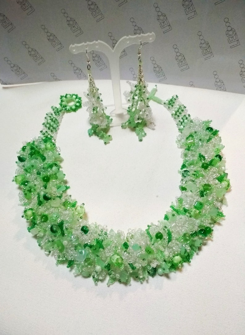 May include: A beaded necklace and matching earrings set. The jewelry features a mix of clear and various shades of green beads. The necklace is a collar style, and the earrings dangle. The jewelry is displayed on a white surface.