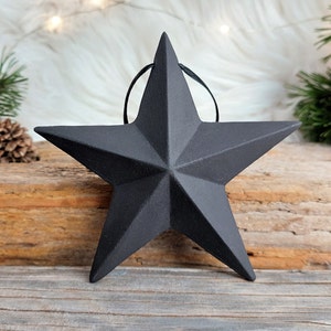 Könnte beinhalten: Ein schwarzer, fünfzackiger Stern-Anhänger mit rustikalem, abgenutztem Finish. Der Stern ist aus Metall und hat eine Schlaufe oben zum Aufhängen.