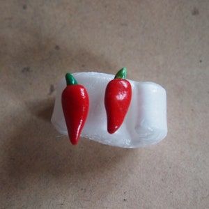 Könnte beinhalten: Ein Paar rote Chili-Pfeffer-Ohrringe mit grünen Stielen, die auf einer weißen Oberfläche liegen. Die Ohrringe sind klein und detailliert, mit glänzendem Finish. Der Hintergrund ist braun.
