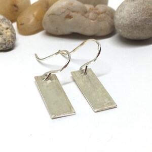 May include: A pair of silver dangle earrings with rectangular shaped charms. The charms have a brushed finish.