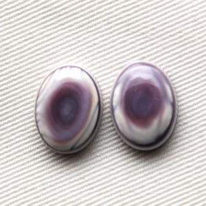 Op de afbeelding: Twee ovale cabochons met een wervelend patroon van paars en wit. De gepolijste stenen hebben een glanzende afwerking en liggen op een lichtgekleurde, getextureerde achtergrond. Geschikt voor het maken van sieraden.
