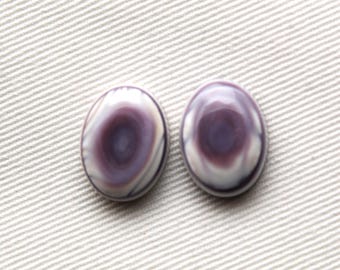 May include: Two oval-shaped cabochons with a swirling pattern of purple and white. The polished stones have a glossy finish and are set against a light-colored, textured background. These could be used for jewelry making.