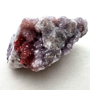 May include: A raw mineral specimen featuring a cluster of crystalline structures. The stone displays a prominent deep red section on the left side, transitioning into lighter shades of purple, lavender, and grey across the textured surface.