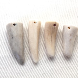 May include: Four individual, tapered, off-white and light grey horn-shaped pendants arranged in a row. Each piece features a smooth, organic texture with a small, circular hole drilled near the wider top end for stringing or attachment purposes.