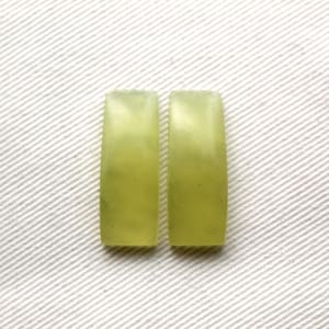 以下が含まれることがあります： 長方形の薄緑色の翡翠の石が2つ。石はわずかに湾曲した形状で、滑らかで研磨された表面を持っています。白いテクスチャード加工された背景に配置されています。